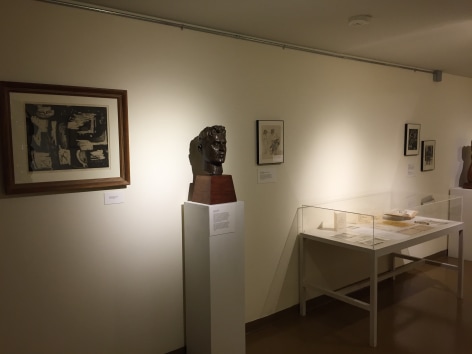White gallery wall going diagonally to the right with medium brown flooring beneath it and a low white ceiling. On the left closest to us is a black and white large artwork with a medium brown frame of various organic shapes. To the right of this piece is a dark brown bust of a man, to the right of this sculpture is a large glass box on white table pieces of paper and a book inside. There is also a small black and white drawing directly to the right of the bust and two small artworks in black frames hung up to the right of the glass box.