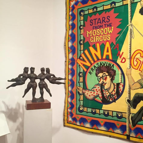 A white gallery wall with a small black sculpture by Chaim Gross, &quot;The Flying Ballerinas&quot; which is a symmetrical composition of four female figures, one standing and holding the other as they fly horizontally outwards on each side on the right, and to the right the left half of a colorful tapestry with the wording &quot;Stars from the Moscow Circus&quot; and a woman in circle frame gesturing to the left on it.