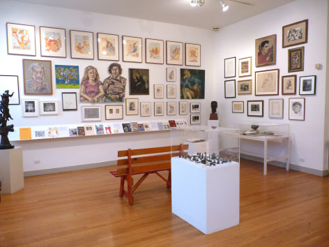 View of two white gallery wall intersection to the center right of us. The walls are crowded with framed art pieces, most prominently a large painting of a female and male figure on the left and a portrait of a young man with dark hair on the top right. Directly in front of us is series of tiny dark colored sculptures mounted on a rectangular box with a glass covering them. There is a light brown wood bench directly behind this installation.