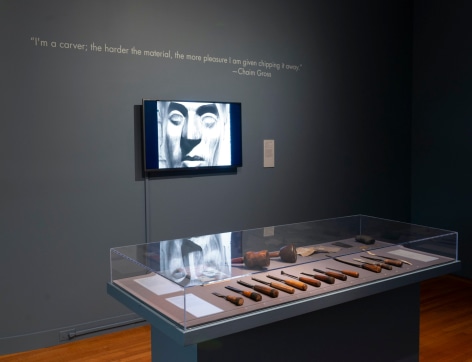 Photo of the exhibit, featuring selected work tools encased in glass, as well as a mounted tv playing a video on Chaim Gross' work. On the wall where the mounted tv is, there is a quote from Chaim Gross in white against the faded navy walls.