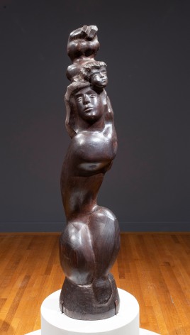 Close up image of Photo of I LOVE MY BABY, 1948, which is positioned on a rounded white base. The wooden sculpture depicts a woman with long hair, crouched so that her legs are drawn close to her tall torso. She is supporting the figure of a baby, whose head touches hers but whose body is raised above its head.