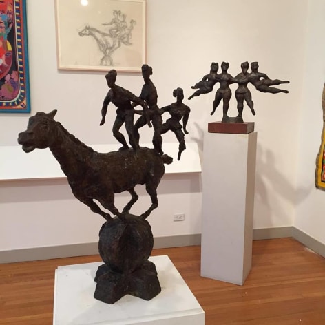 Photo of a white gallery wall with light brown wood flooring beneath it. In front of us is a large black sculpture of three tiny people balancing on a gigantic horse. Behind this sculpture is another black sculpture by Chaim Gross, &quot;The Flying Ballerinas&quot; which is a symmetrical composition of four female figures, one standing and holding the other as they fly horizontally outwards on each side. On the wall behind the sculptures is a large line drawing of the horse sculpture in a light cream frame. Next to the drawing the corner of a large, colorful artwork is barely visible.