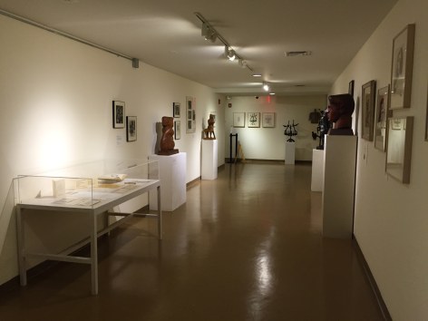 Hallway of a gallery with white walls, medium brown floors, and a low white ceiling. On the left wall closest to us is a glass box mounted on a white table with papers and a book inside. Next to it on the left wall towards the right is a medium brown, mid sized sculpture of a vaguely humanoid, organic shape, and to the right of that piece is a unidentifiable animal like, organic form. There are four medium sized framed artworks and one large piece between and around these sculptures. On the right wall there is a large, medium brown bust of a man and a piece of a black unidentifiable sculpture is visible. Closest to us on the right are the sides of four artworks in beige frames. On the back wall there is a black sculpture of a tree with birds on it and three mid sized artworks hung up to the left of it.