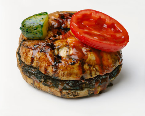 Photograph by Sharon Core of a hamburger covered in sauce with pickle and tomato on top of bun arranged to look like the sculpture by Oldenburg