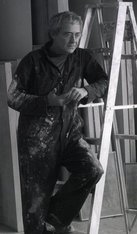 A black and white photos of Robert Natkin wearing paint-splattered overalls leaning against a ladder.