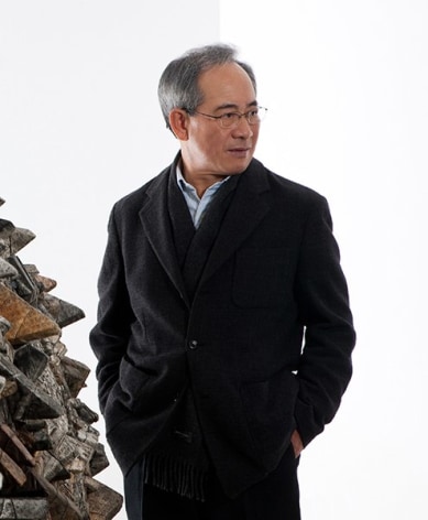 Portrait of Artist Chun Kwang Young wearing a black jacket, standing next an installation composed of foam triangles covered in antique pages of Korean  books.