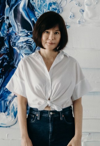 Portrait of artist Jane Lee wearing a shite shirt and Jeans standing in front of a textured white and blue painting.
