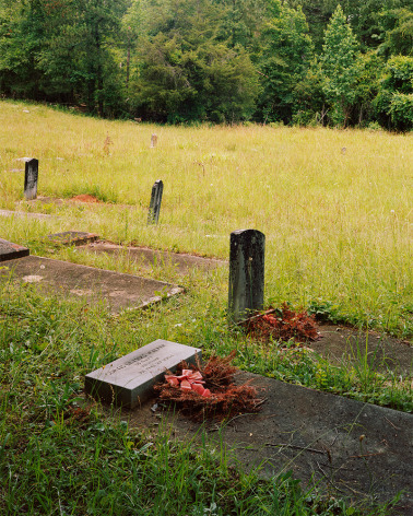 Vertical photograph of headstones in a cemetery, green grass, with dark green trees in the distance, wreathes of red and orange flowers at the gravesites