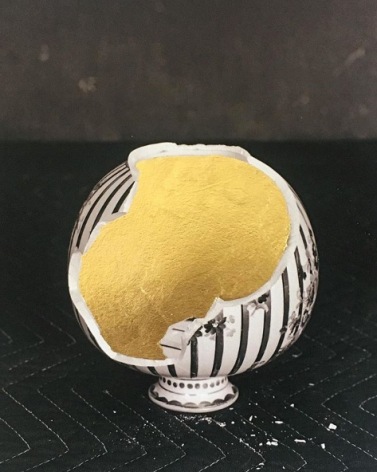 James Henkel  Repaired Upside Down Vase, 2019  Archival Pigment Print with Gold Leaf  10h x 8w in , Black and white photograph of a broken, round, striped, vase, upside down on a black blanket. With hand painted gold leaf filling the vase center.