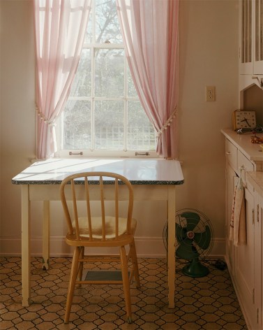 Vertical photograph of a 1950s style kitchen with a small table and one chair underneath a window with sheer pink curtains. A metal fan sits on the worm linoleum floor