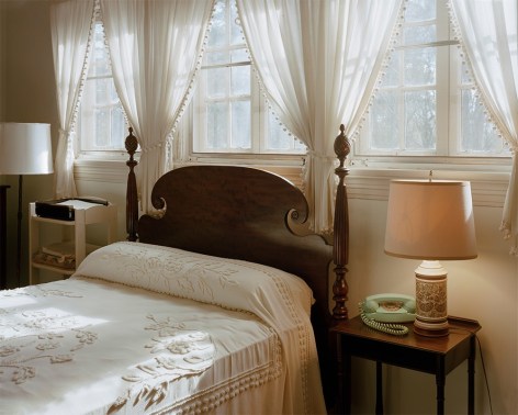 horizontal photograph of a upper middle class bedroom. A poster bed sits underneath a trio of windows with decorative curtains. A writing table and nightstand with lamp sit to the right and left.