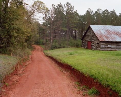 Horizontal photograph of a red clay dirt road leading into the woods. there is a strip of green grass to the right with a wooden barn structure with a tin roof and a red door