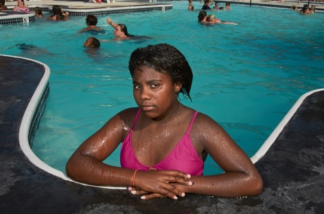 photograph of a young black girl in a bright pink swimsuit staring at the camera from a pool