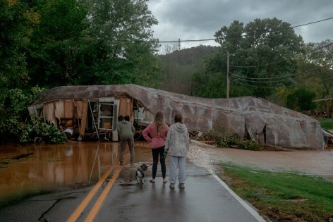 three people and a dog stand on a flooded road looking towards a destroyed trailer