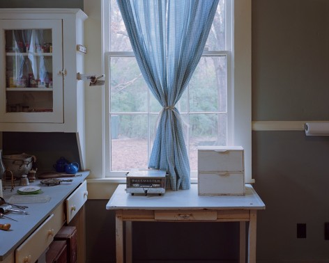 Horizontal photograph of Willaim Faulkner's kitchen at Rowan Oak. A blue curtain is in the middle of the window, with a small table underneath. It seems a typical southern kitchen