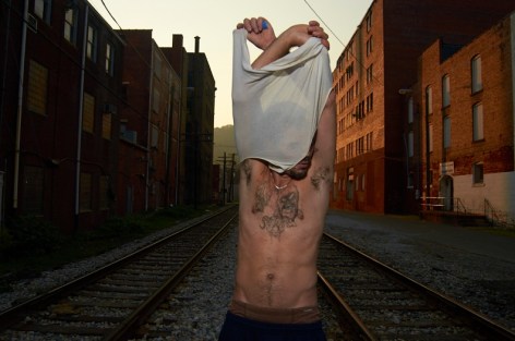Stacy Kranitz  Logan, West Virginia, 2011  Archival Pigment Print  16h x 24w inches, Edition of 5  30h x 40w inches, Edition of 3, exterior scene of a man in the middle of the street removing his white t-shirt revealing his bare chest. Brick building along both sides of the street