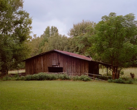 horizontal photograph of a wooden horse barn with a red roof in the middle of a green pasture. There are various tress in the background