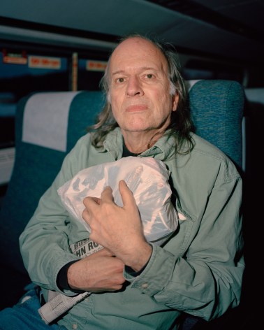 Older person holding plastic bag and newspaper on train, by McNair Evans