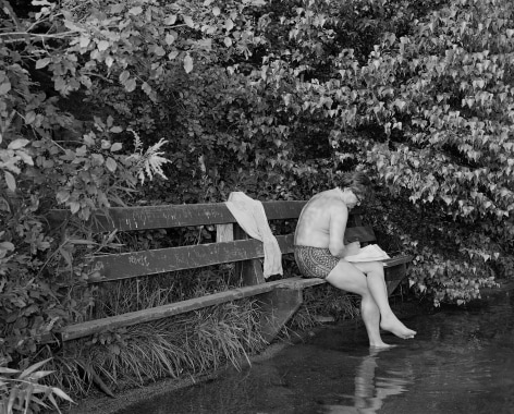 1970s black and white photograph of person on park bench with back turned and feel in water, by Mike Smith