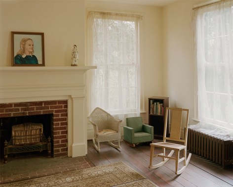 Photograph of a historic bedroom of the daughter of William Faulkner. A portrait of the young girl sits on a mantle able the fireplace, two rockers, book case and a green chair are bu the window