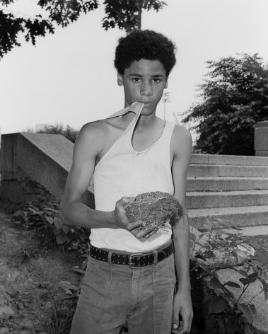 Boy holding a bird's nest and biting his tank top in a park. Photograph by Mike Smith