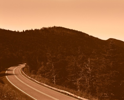 Warm hued landscape image of two lane road through mountain terrain, by Gesche Wurfel