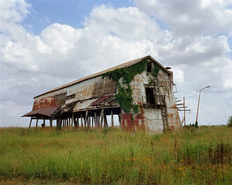 Horizontal photograph of a decaying cotton gin building. There are multiple layers of peeling paint and ivy taking over the facade.