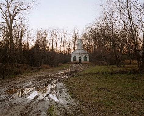 Horizontal photograph a muddy dirt road leading back to a white wood slat church in the woods