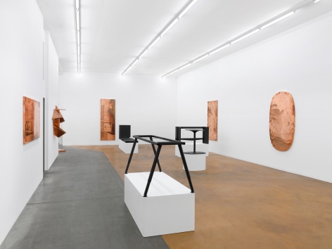 Installation view of Beshty's exhibition at MAMCO featuring several copper surrogates on the walls with table and desks without tops on pedestals in the center of the room.