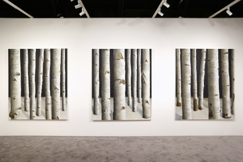 Three of the same sized paintings of a forest of trees with text scrawled into the bark is shown hanging side by side in an art fair booth.
