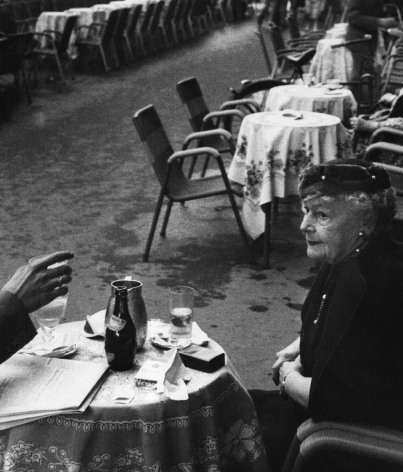 Piergiorgio Branzi, Roma, Via Veneto, ​1959. An older woman seated at the foremost of a row of tables. A hand holding a glass is extended towards her from outside the left of the frame.