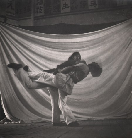 Cecil Beaton, Chinese College of Physical Training, ​1945. Two figures blurred with motion in front of a white cloth backdrop. One leans forward and the other back, each with one leg raised, seemingly in a fighting match.