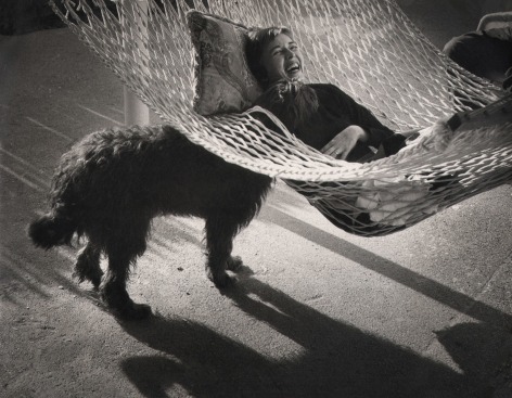 John Bryson, A 'family' picture of the photographer's wife and dog, ​c. 1959. A woman reclines in a hammock in the upper right of the frame, a black dog stands beneath the hammock to the left, poking its head through the hammock to the woman's face.