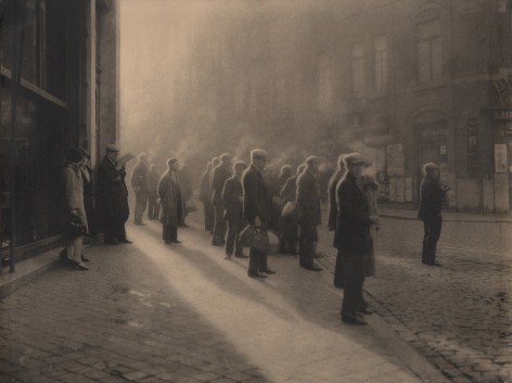 08. L&eacute;onard Misonne, Premi&egrave;re cigarette, 1927. Various figures, mostly capped men, stand on the sidewalk, cigarette smoke rising above them. Sepia-toned print.
