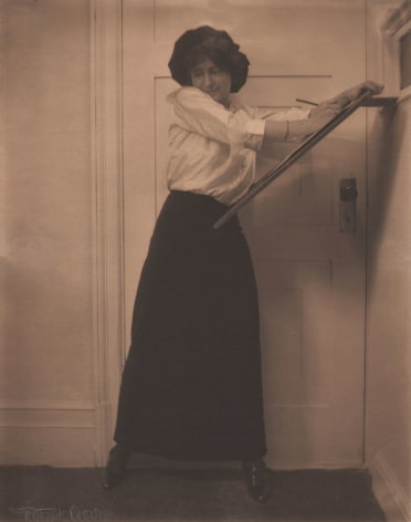 01. Gertrude K&auml;sebier, Untitled, ​c. 1910. A woman in a white shirt and long black skirt standing in front of a door, seeming to draw against a wooden easel.