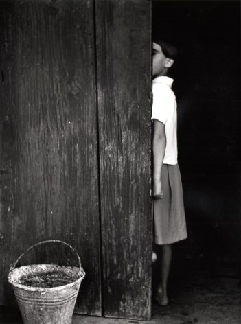 Nino Migliori, People of Emilia, 1950. A young girl is half hidden behind a wooden door. A bucket is in the lower left of the frame.