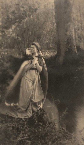 07. Dorothy &amp; Reta Morter, Destiny - Self-Portrait, ​c. 1918. A woman stands in the left of the frame by a river in a dress and cloak, looking to the left.
