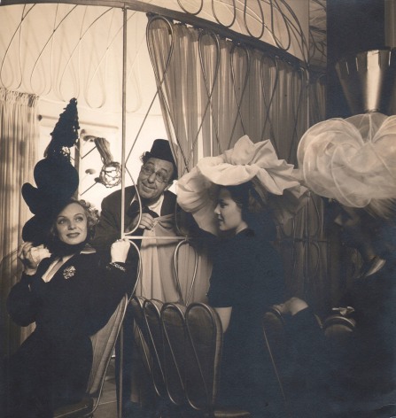 Louise Dahl-Wolfe, Ed Wynn &amp; Jane Pickens In Boys &amp; Girls Together, 1940. A man in glasses and a top hat peers out from behind a curtain. Three women in large billowing hats are seated in the foreground.