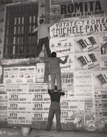 Federico Garolla, Elezione a Napoli, ​1948. Three men stand on each others shoulders to reach a high point on a wall covered in election posters.