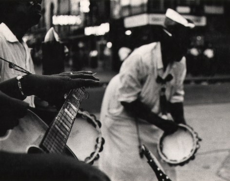 04. Beuford Smith, Sunday in Harlem, ​1968. Figures playing tambourines and a guitar on the street. Focus is on the lower left of the frame on the neck of a guitar and the player's left hand.