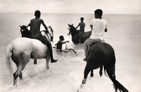 04. Mike Andrews, Nassau, Bahamas. Stable boys exercising racehorses on Cable Beach, c. 1960. Four boys on horses in knee-deep water. A fifth boy is in the water with a bicycle.