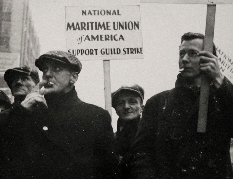 Gordon Coster, W.P.A. Parade, Chicago, 1939. Four men walk in black coats on a snowy day, smoking cigarettes. One holds a sign that reads &quot;National Maritime Union of America Support Guild Strike&quot;