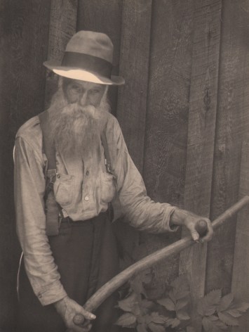 10. Doris Ulmann, Untitled (Farmer holding oxen yoke), 1928&ndash;1934. Older bearded man in a hat standing against a wooden wall and suspenders holding a wooden rod with two handles.