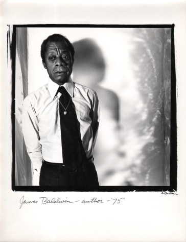 Anthony Barboza, James Baldwin, Author, ​1975. Subject stands to the left side of a square frame, looking down and to the left. His shadow is cast to the right on the wall behind him.