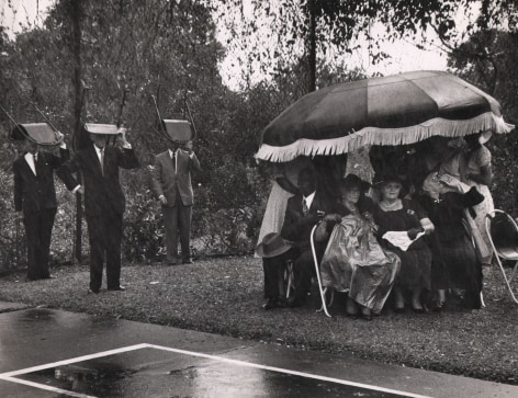 Leonard McCome, Governor's Garden Party, Grenada Island, ​c. 1959. Guests huddle under a large umbrella on the right of the frame while three suited men on the left hold chairs above their heads.