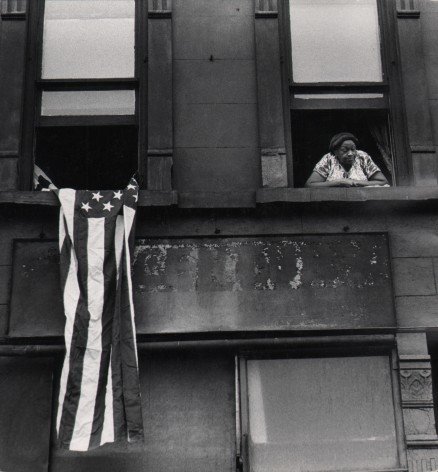 01. Beuford Smith, Flag Day, Harlem, ​1976. Detail of two windows of an apartment building; a woman leans out of the right window, an American flag hangs from the left.