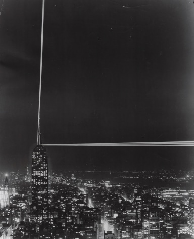 08. Anonymous, Patterns in Light: the Empire State's controversial beacons, 1956. Distant, head-on nighttime view of the Empire State Building (lower left of frame), with two beams of light streaming up and to the right.
