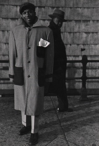 Marvin E. Newman, Chicago, ​1950. A young adult male in a large coat stands on the sidewalk, smiling to the camera, another man in black walks by in the background.