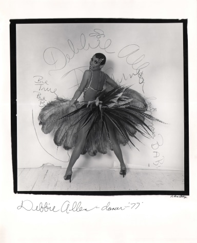 Anthony Barboza, Debbie Allen - Dancer, ​1977. Subject stands in feathered skirt, back to camera, legs wide. Head is turned smiling to camera. Writing on white backdrop includes her name.