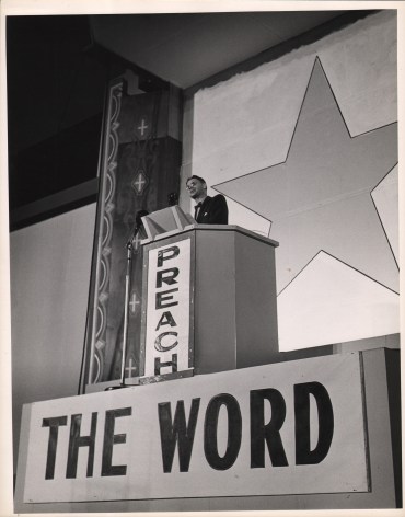 Gordon Coster, Preach the Word, ​c. 1940. A suited man stands at a raised podium that reads &quot;Preach The Word&quot;. A large star is painted on the wall behind him.